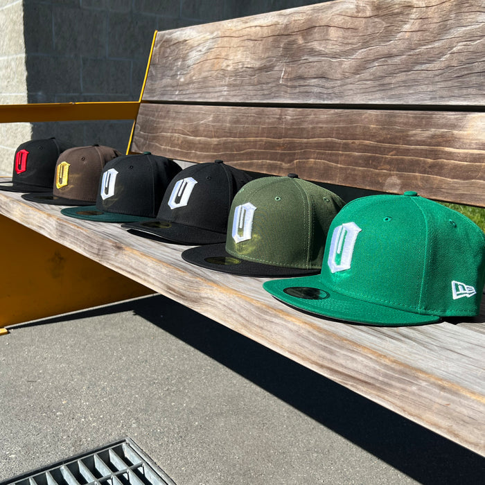 Six New Era Official 59FIFTY Fitted Caps by Other—in black, brown, olive, and green—each featuring a bold embroidered letter “D” on the front in classic fitted style, are neatly arranged on a wooden bench outdoors, with sunlight accentuating their colors.