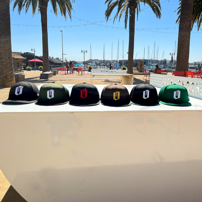 Six New Era Official 59FIFTY Fitted Caps from Other are shown in classic black and green styles, each featuring a stylized “O” logo in white, red, gold, or black. The caps are lined up on a white surface outdoors with palm trees, string lights, red chairs, and a marina with docked boats visible in the background.