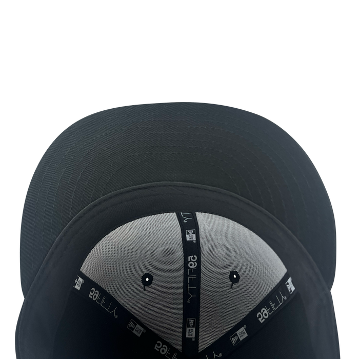 A black New Era Pericos ALT 59FIFTY Cap viewed from underneath, displaying the brim and interior panels. The inner seams are finished with black taping featuring white symbols and text typical of New Era caps, while two small ventilation holes can be seen on the grey fabric. The background is plain white.