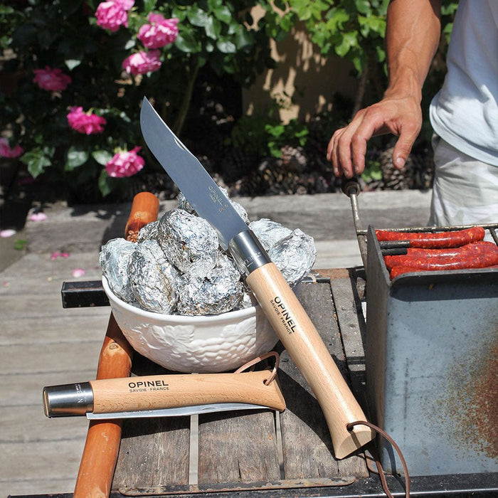 A giant Opinel knife leaning on bowl of aluminum foil-covered foods on outdoor grill.