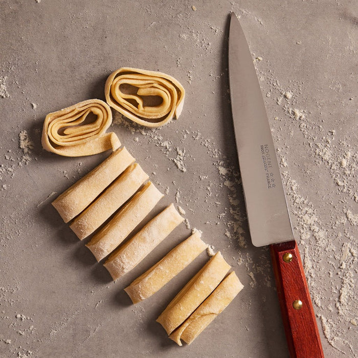 Kitchen knife with wood handle next to raw pasta on floured gray surface.