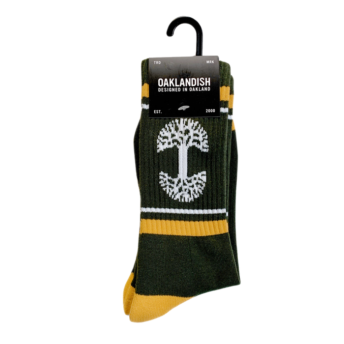 Introducing the OAK Knit Crew Socks by Oaklandish, a stylish pair crafted in forest green with yellow accents and adorned with a white tree design at the center. Made from a comfortable blend of cotton, polyester, and spandex, these crew socks also showcase the Oaklandish logo. They come packaged with a sleek black label that reads "OAKLANDISH - Designed in Oakland - Est. 2000," and include a convenient plastic hanger for easy display.