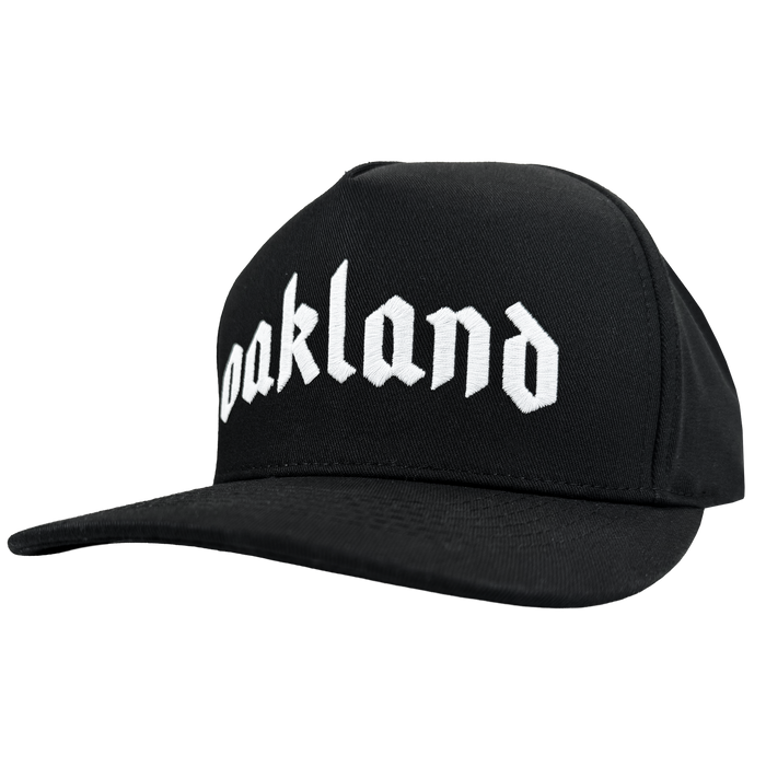 The Oaklandish Oakland 68s Panel Cap is a black snapback with "Oakland" boldly embroidered in white Old English style on the front. It features a flat brim and is displayed against a plain, light background.