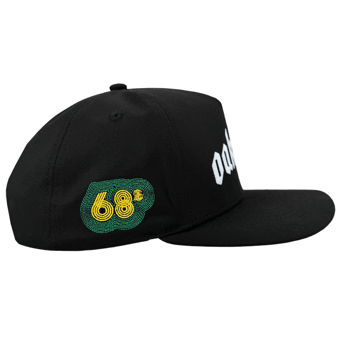 The Oaklandish Oakland 68s Panel Cap is a black snapback hat with a flat brim, structured crown, green and yellow embroidered “68” on the left panel, partially visible white text on the front, shown from the side on a white background.
