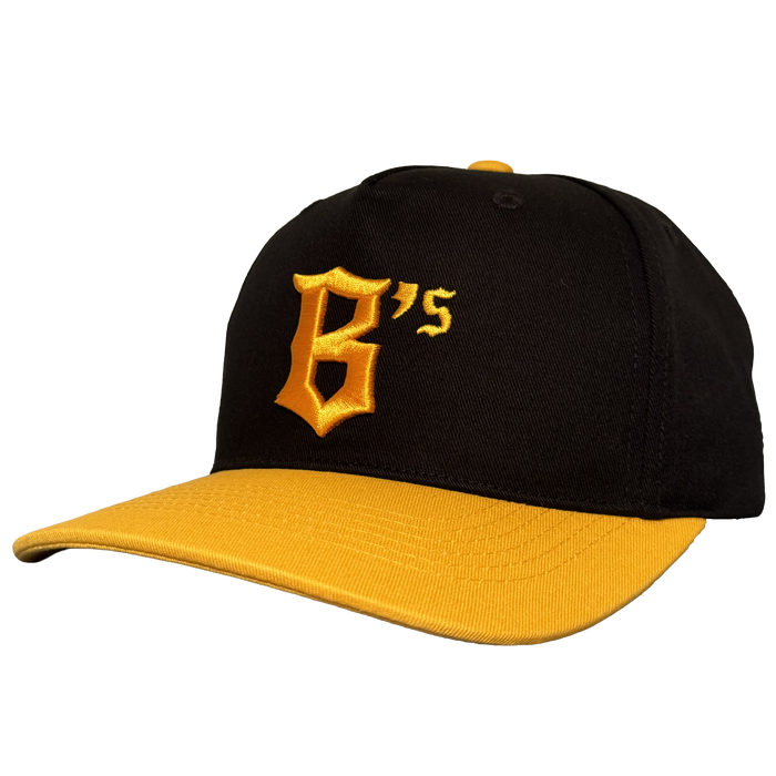 The Oakland Ballers 5 Panel Cap by Oakland Ballers features a black snapback design with a yellow brim and button, showcasing a bold yellow "B’s" stitched on the front left, inspired by the team. The cap is angled left on a plain white background.