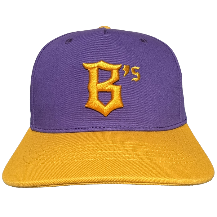The Oakland Ballers 5 Panel Cap by Oakland Ballers features a stylish purple snapback design with a flat gold brim. On the front, it showcases a prominent gold letter “B” with an embroidered gold “’s” in the top right corner, both in a distinctive font that pays homage to the Oakland Ballers. The cap is finished with two small black ventilation holes on top, blending comfort and style—perfect for any fan of Oakland’s baseball team.