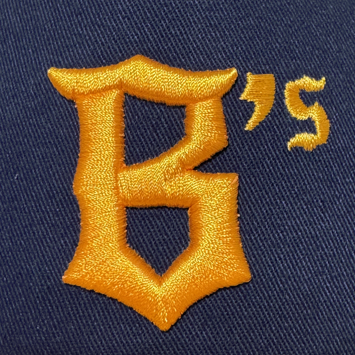 An embroidered gold "B" with an old-fashioned style, apostrophe, and "S" appears on dark blue twill fabric of the Oakland Ballers 5 Panel Cap by Oakland Ballers. Detailed stitching suggests it’s part of their snapback hat collection.