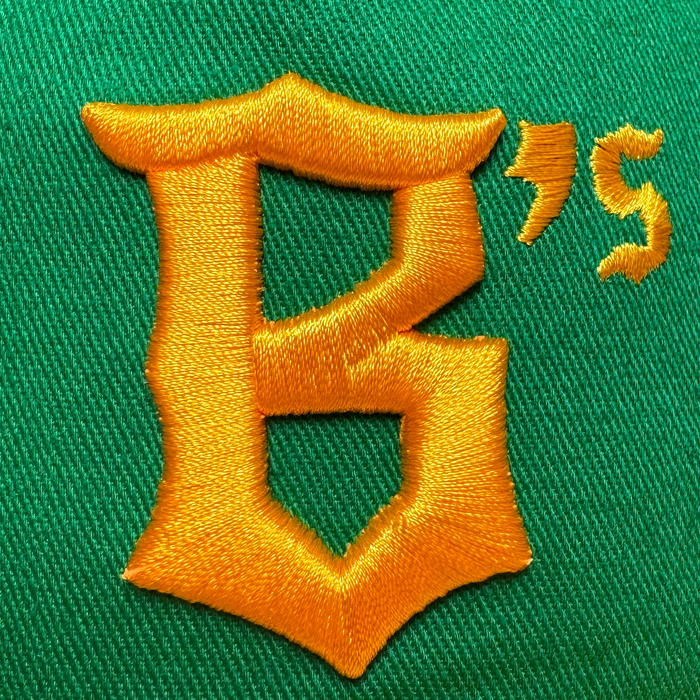 Golden yellow embroidered "B" and "S" letters in bold, stylized font on green fabric appear on the Oakland Ballers 5 Panel Cap by Oakland Ballers, with visible stitching highlighting its connection to the Oakland baseball team.