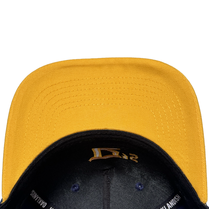 A close-up, top-down view of the Oakland Ballers 5 Panel Cap by Oakland Ballers shows its yellow color, black underside, brim stitching, black embroidered interior details, and partial white text on the inner band against a white background.