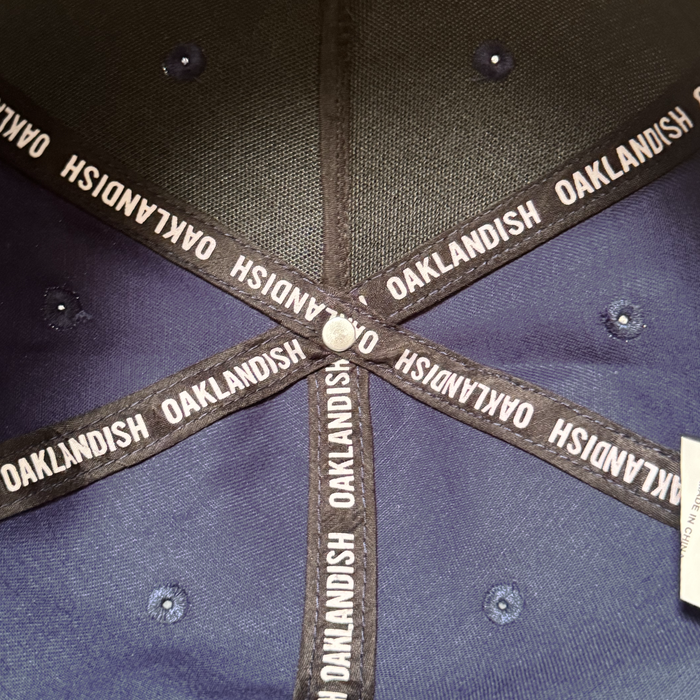 Close-up of the Oakland Ballers 5 Panel Cap’s navy blue interior. Black “OAKLANDISH” bands cross below a metallic button, with blue stitched eyelets and a white tag—ideal for Oakland Ballers or Bay Area baseball fans.