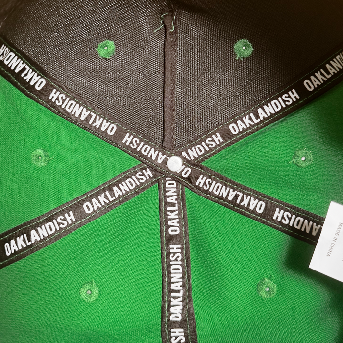 Close-up of the inside top of the Oakland Ballers 5 Panel Cap. Black fabric strips with repeated white “OAKLANDISH” text form an X at a white button, while green stitched holes and a white tag reflect classic Oakland baseball style. Brand: Oakland Ballers.