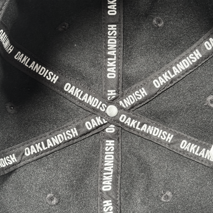 Close-up of the inside of an Oakland Ballers B Strapback hat, showing crossed black fabric strips with “OAKLANDISH” in white uppercase letters and a metal button at the intersection. Brand: Oakland Ballers.
