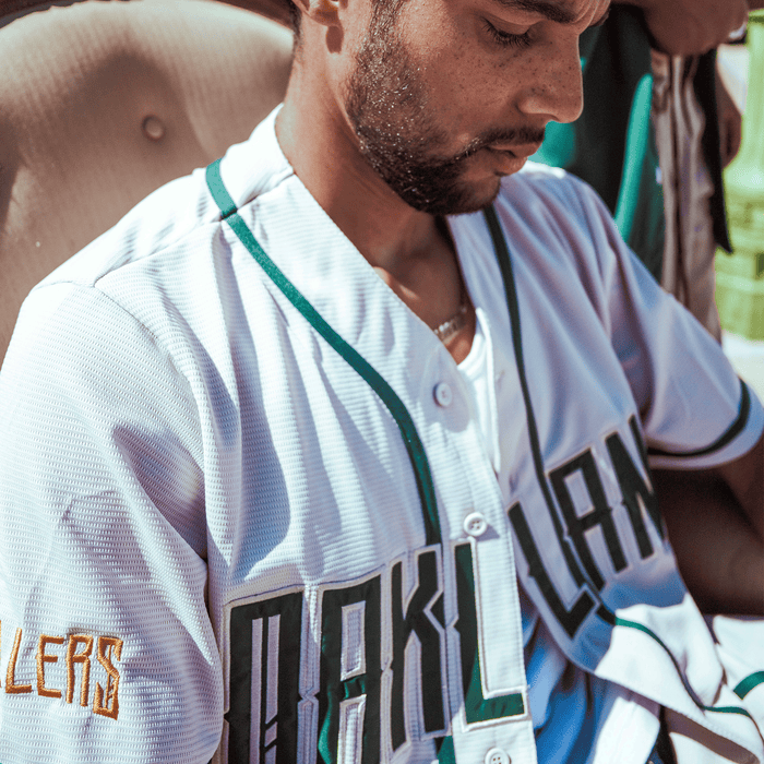 A man in an Oakland Ballers Button Up Jersey, featuring white fabric with green trim and yellow accents, sits looking down. The word “HUSTLERS” appears on his sleeve as sunlight casts shadows across his focused face.