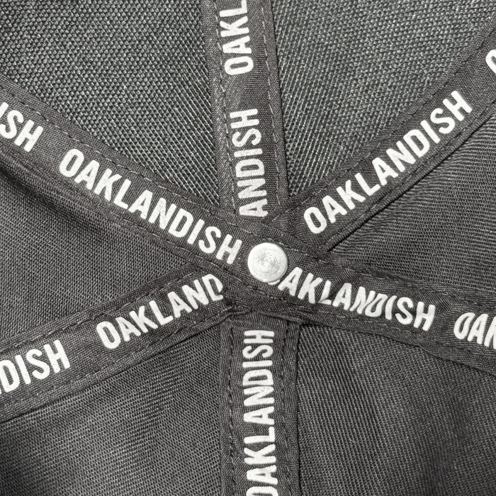 Close-up of the inside of an Oakland Ballers Flexfit Cap, showing intersecting black fabric bands with "OAKLANDISH" in white, a small silver center button, and mesh polyester fabric in the background. Brand: Oakland Ballers.