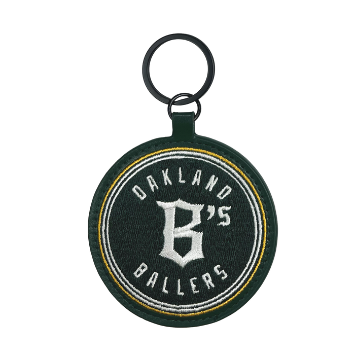 The Oakland Ballers Keychain features a round, dark green fabric embroidered with "Oakland Ballers" in white and a large "B's" at the center. It includes a black ring at the top for attaching keys and has a green border surrounding the entire design. The text is highlighted with yellow and white accent lines, celebrating this pioneering professional baseball team.