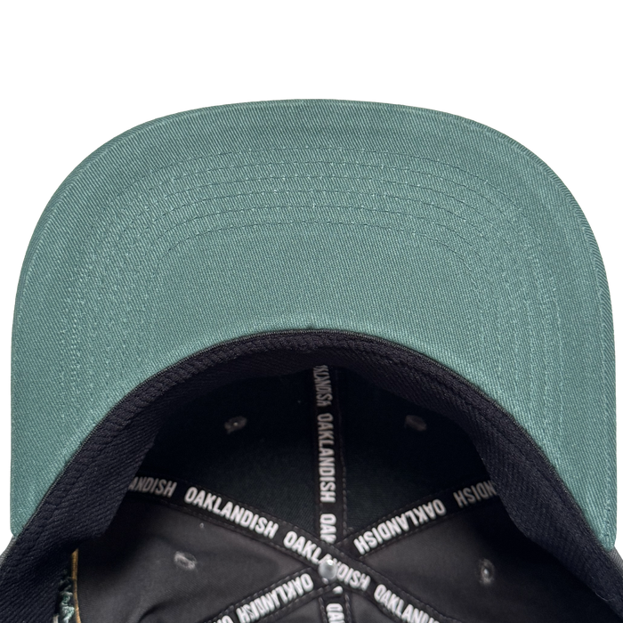 A top-down view of the Oakland Ballers Logo Hat by Oakland Ballers shows the green cap with black brim, white “OAKLANDISH” text on the inner band, and metal rivets around the crown, celebrating the Oakland baseball team.