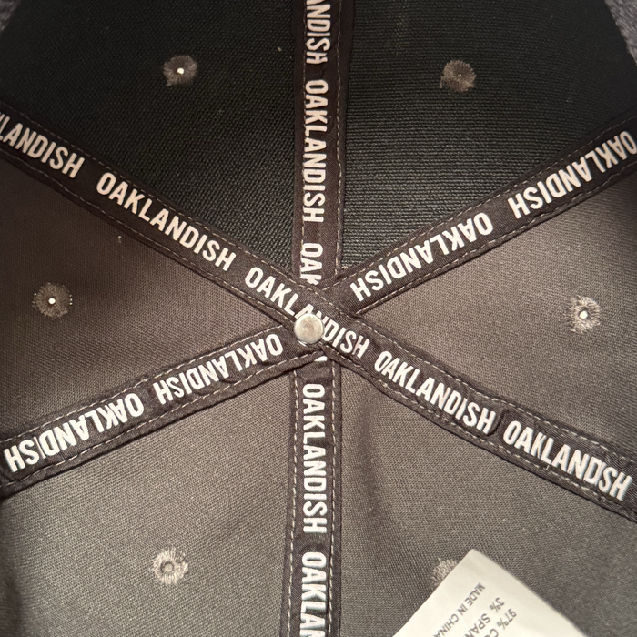 Close-up of the Oakland Ballers Logo Hat’s black interior shows fabric strips with repeating white "OAKLANDISH" text forming a star around a metal button, referencing the Pioneer League. Stitched ventilation holes and a white tag are also visible.