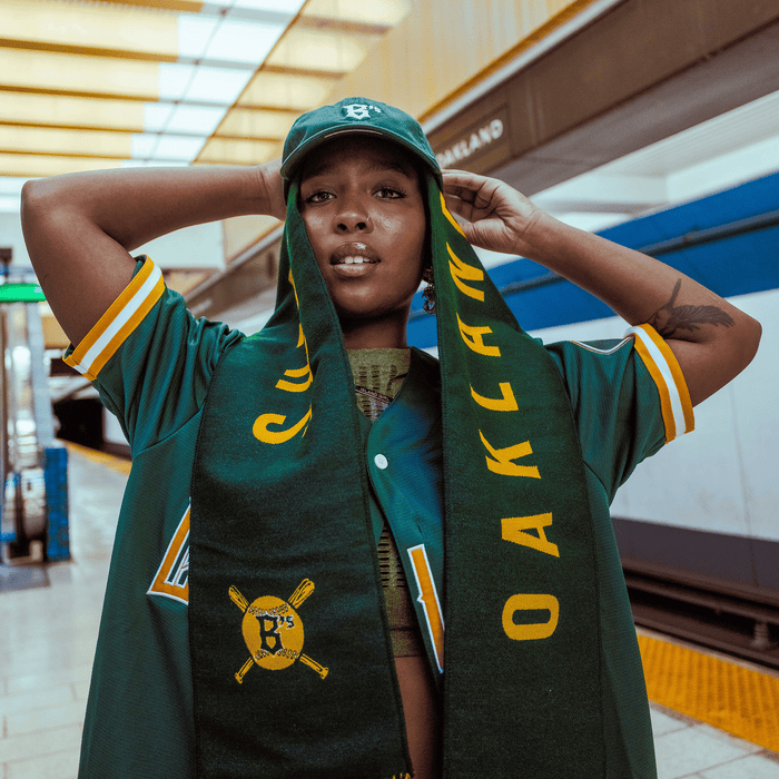 A woman on a subway platform confidently holds up an Oakland Ballers Scarf (by Oakland Ballers) in green and yellow. She wears matching team gear, with a train behind her and "OAKLAND" visible on the station sign.