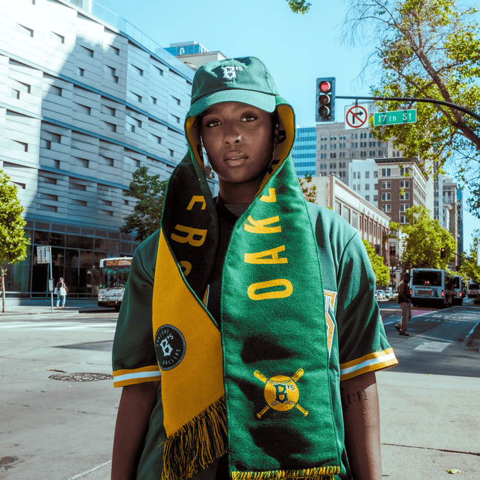 A person stands on a city street in Oakland Ballers gear, including a green jersey, bucket hat, and an Oakland Ballers Scarf. A “17th St” sign highlights the home of Oakland’s pro teams, including the new Pioneer League club.