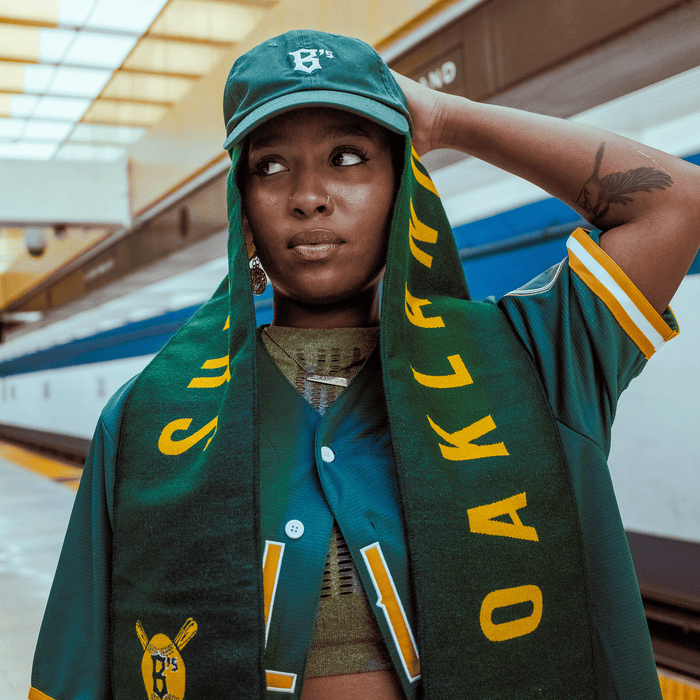 A woman at a train platform, proudly wearing an Oakland Ballers Scarf by Oakland Ballers, a green cap, and jersey, shows her city pride—celebrating the new Pioneer League pro baseball team: the Oakland Ballers.