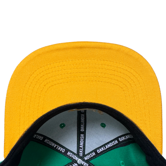 A close-up, top-down view of the Oakland Ballers Snapback by Oakland Ballers features a yellow cap with green interior, black brim underside, and an "OAKLANDISH" inner band, echoing Oakland baseball style on a white background.