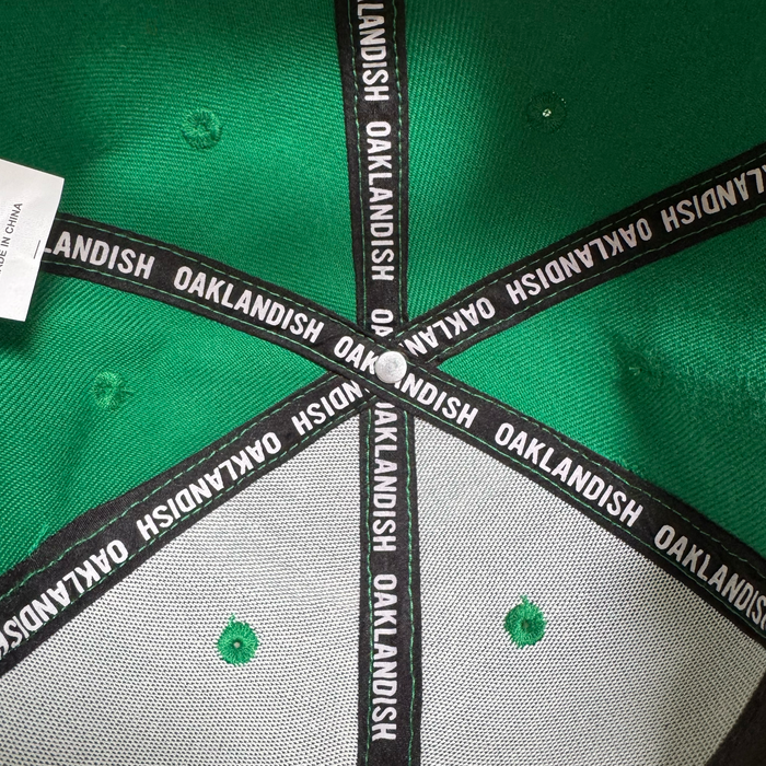 Close-up of the inside top of an Oakland Ballers Snapback by Oakland Ballers. Black fabric strips labeled “OAKLANDISH” crisscross above green stitching and ventilation holes, celebrating Oakland baseball team spirit.