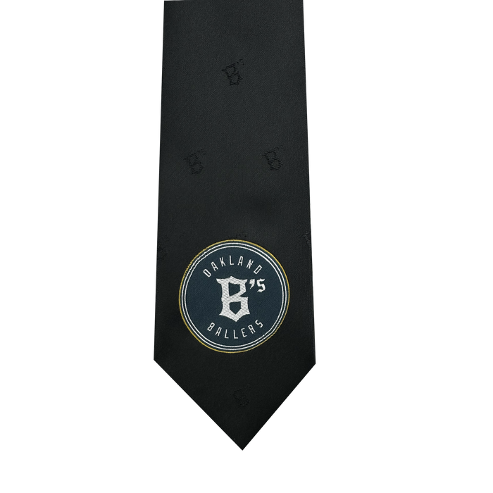 Introducing the Oakland Ballers Tie by the renowned brand Oakland Ballers. This elegant black tie features a subtle pattern and an eye-catching embroidered patch near the bottom. The patch proudly displays "Oakland Ballers 75" alongside a stylized "B," honoring the professional baseball team from the Pioneer League. The design is showcased in white text on a dark circular backdrop, elegantly outlined in gold.