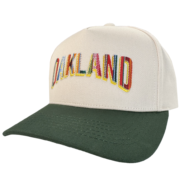 The Oakland Beisbol 5 Panel Cap by Oakland Ballers features a beige pro baseball snapback, dark green brim, and "OAKLAND" in colorful letters across the front—celebrating the team. Displayed angled to show the front panel; white background.