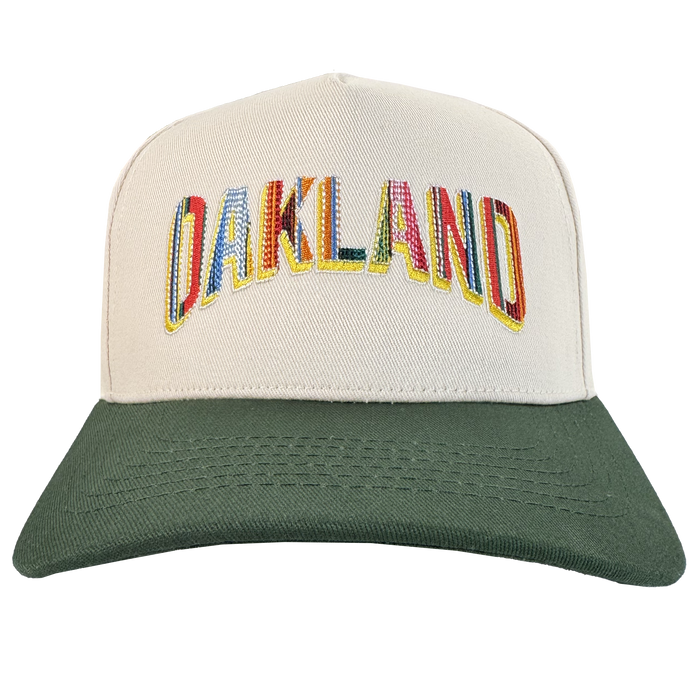 The Oakland Ballers Oakland Beisbol 5 Panel Cap features a beige body, dark green brim, and "OAKLAND" in large, patterned letters outlined in yellow—celebrating Oakland baseball's vibrant spirit. Photo background is plain white.