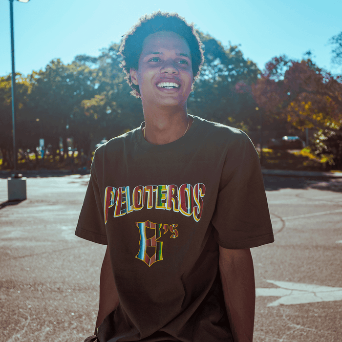 A smiling young person with short curly hair wears the Oakland Ballers Oakland Beisbol Tee outdoors, with autumn trees and a sunny sky behind them, reflecting the energetic spirit of the Pioneer League baseball team.