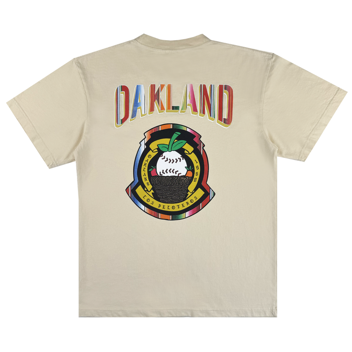 The Oakland Beisbol Tee by Oakland Ballers is a beige shirt featuring "OAKLAND" in bright letters on the back, plus a vibrant emblem with a baseball, green leaves, and "POR LOS PELOTEROS" on yellow and red banners.