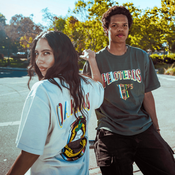 Two young adults pose outside under blue sky and trees. She wears a white tee with a colorful graphic; he sports the Oakland Ballers "Oakland Beisbol Tee," repping Oakland’s Pioneer League baseball team.