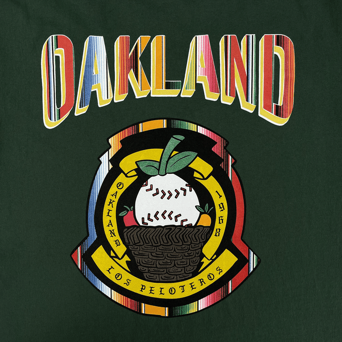 The Oakland Beisbol Tee by Oakland Ballers is a green shirt with "OAKLAND" in bold, colorful letters above a circular patch featuring a baseball plant in a basket and the text "OAKLAND - 1968 - LOS PELOTEROS.