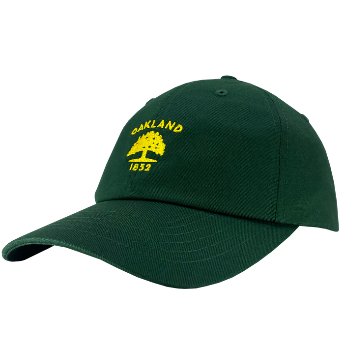 The Oaklandish "Oakland Flag Dad Hat" is a dark green cap adorned with yellow embroidery on the front. The design features the text "OAKLAND" in an arch above an illustrated tree, reminiscent of the Oakland flag, with "1852" below. The hat has a curved brim and an adjustable strap at the back.