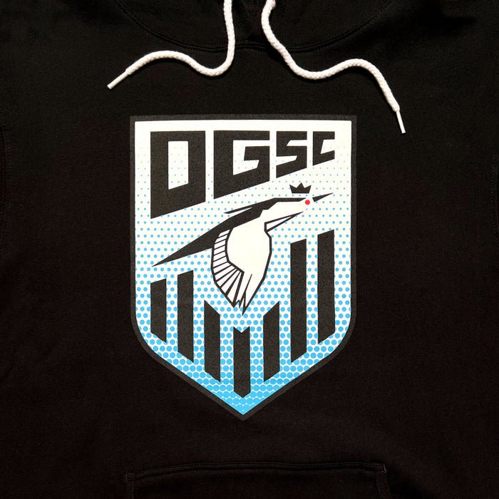 The image displays the Oakland Genesis Heron Hoodie from Partner. It is a black hoodie adorned with a crest-style logo that showcases a stylized bird featuring a red eye and a crown above its head, along with vertical blue stripes below. "Oakland Genesis" and the letters "DGSE" are prominently positioned at the top of the logo in bold, angular lettering. The hoodie also includes white drawstrings for added detail.