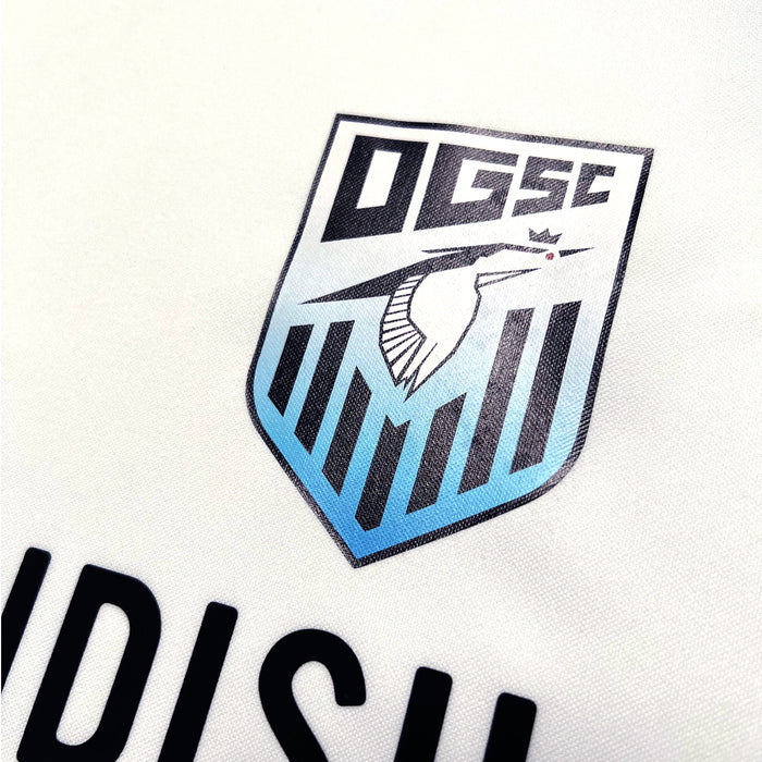 Close-up image of the Oakland Genesis Home White jersey by Partner. The youth soccer team's logo features a stylized kingfisher with a crown on its head, placed inside a shield adorned with vertical stripes. The letters "OGSC," symbolizing Oakland Genesis, are prominently displayed at the top of the shield.