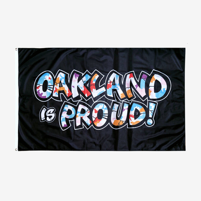 Introducing the Oaklandish "Oakland is Proud Flag" – a black flag with the words "OAKLAND is PROUD!" displayed in bold, colorful graffiti-style letters, evoking the essence of an iconic Oakland mural. The word "OAKLAND" bursts with vibrant colors and patterns, while "is PROUD!" features a primarily white aesthetic with color accents. Metal grommets on the left side facilitate easy hanging as an accessory.