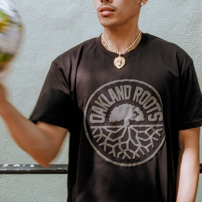 Wearing the Oakland Roots SC Blackout Logo Tee by Oakland Roots SC, a person stands against a light wall. They spin a blurred soccer ball in one hand, sport a gold chain necklace, and their face is partially out of frame.