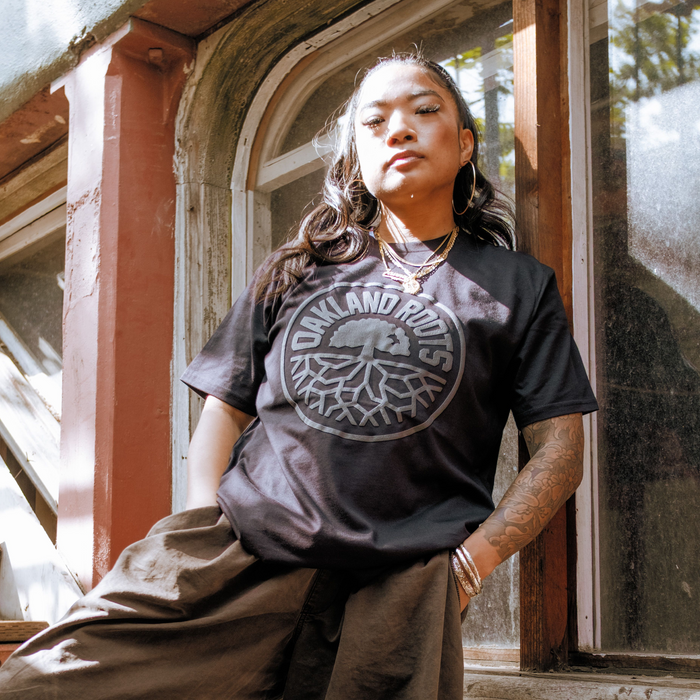 A person with long dark hair stands confidently by a weathered window, wearing the Oakland Roots SC Blackout Logo Tee and brown loose pants, accessorized with gold jewelry and right arm tattoos as sunlight filters through the trees.