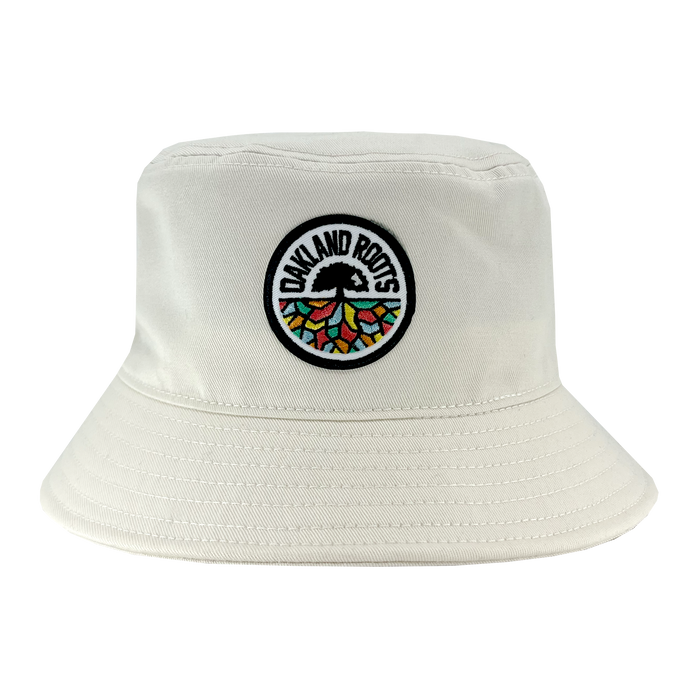The Oakland Roots SC Bucket Hat, by Oakland Roots SC, showcases a light-colored design with an embroidered circular patch on the front. The patch features a tree with colorful segments and roots, encircled by the text "Oakland Roots," celebrating the local sports club. The hat also includes detailed stitching along the brim and crown.
