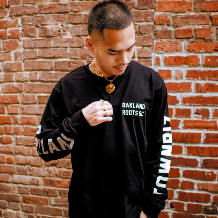 A man with short hair stands against a red brick wall, wearing the Oakland Roots SC Classic 2.0 LS Tee by Oakland Roots SC, featuring "OAKLAND ROOTS SC" on the chest and "TOWN BIZ" on the sleeves. He looks down, touching his shirt.