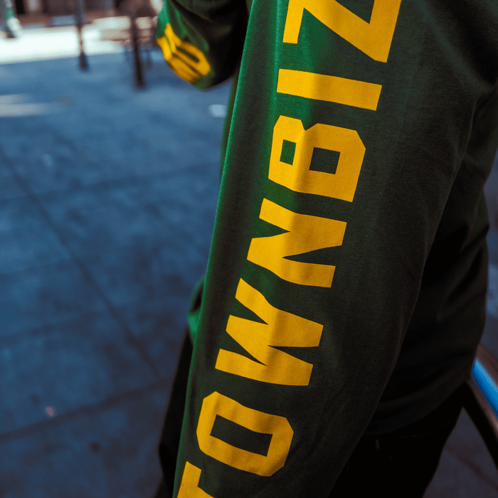 A person wearing an Oakland Roots SC Classic 2.0 LS Tee stands on a sunlit city sidewalk. Only their torso and arm are visible, with a blurred urban background. The tee features bold yellow "TOWNBIZ" lettering down the right sleeve.