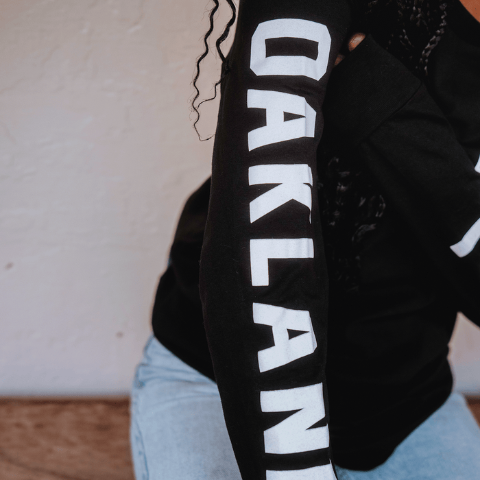 A person wears light blue jeans and a black Oakland Roots SC Classic 2.0 LS Tee with "OAKLAND" in bold white letters down the right sleeve. Their face is unseen as they sit against a textured light wall, embodying community spirit.