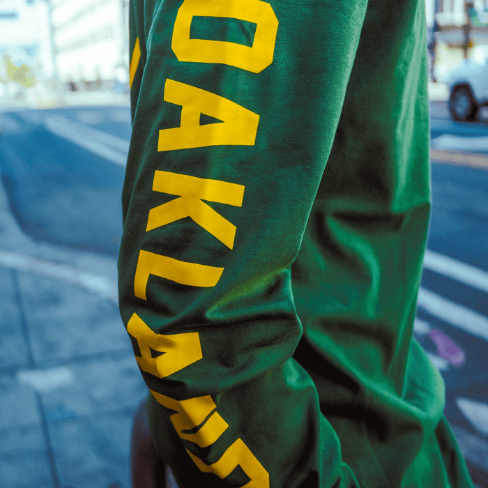 Someone stands on a city sidewalk in an Oakland Roots SC Classic 2.0 LS Tee by Oakland Roots SC, featuring "OAKLAND" in bold yellow letters down the green sleeve, with blurred buildings and cars in the background.