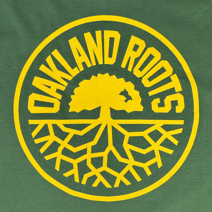 The Oakland Roots SC Classic 2.0 LS Tee by Oakland Roots SC features a circular logo with "OAKLAND ROOTS" in yellow encircling a tree with branches and roots on a green background, symbolizing community ties and social impact.