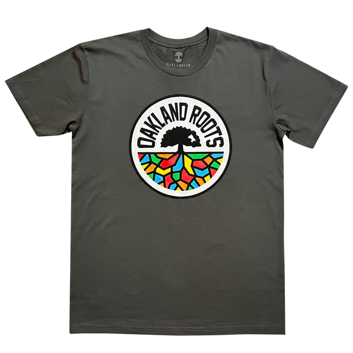 Introducing the Oakland Roots SC Classic Tee by Oakland Roots SC: a charcoal gray T-shirt prominently featuring the "Oakland Roots" logo at its center. The logo, inspired by the Oaklandish aesthetic, showcases a tree with extensive roots forming vibrant mosaic-like patterns in shades of red, blue, green, yellow, and orange. The text "OAKLAND ROOTS" elegantly arches over the tree and its colorful roots.
