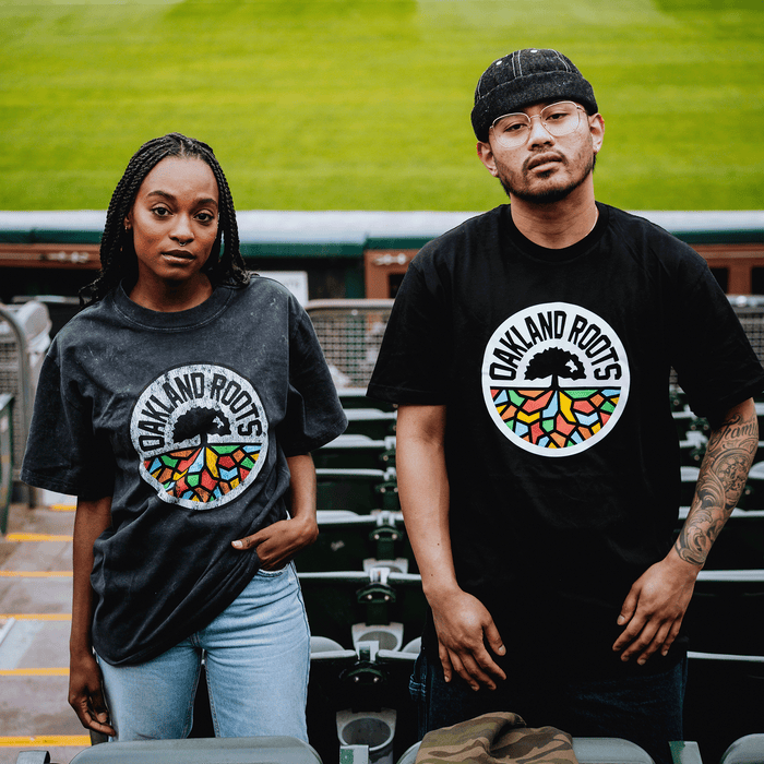 Two people in stadium seating wear black Oakland Roots SC Classic Tees by Oakland Roots SC, each with a colorful tree logo. The woman on the left has long braids; the man on the right wears glasses, a cap, and reveals his tattooed arm.