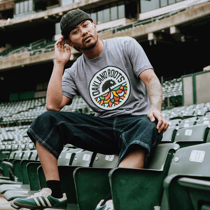 Wearing glasses, a dark knit cap, denim shorts, and sneakers, a man in a gray Oakland Roots SC Classic Tee by Oakland Roots SC sits confidently on stadium seats, touching his cap and reflecting the spirited energy of the team.