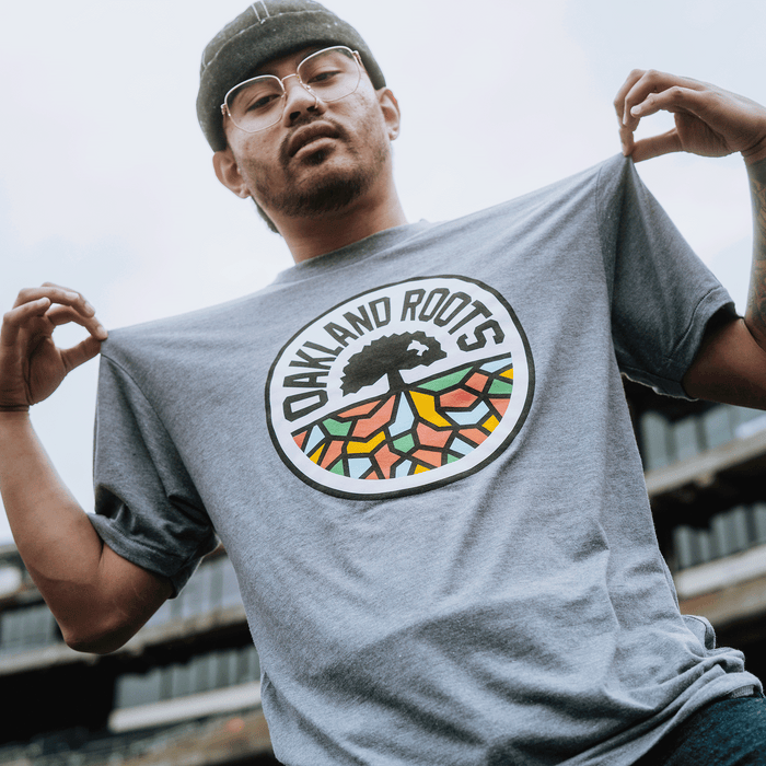 A man wearing glasses and a grey beanie displays the Oakland Roots SC Classic Tee by holding out his shirt, showcasing its colorful round "Oakland Roots Sports Club" logo with tree and roots. He stands outdoors, blurred buildings in the background.