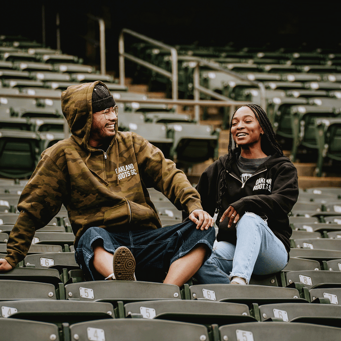 A man and woman smile on stadium seats wearing Oakland Roots SC Classic Zip Hoodies, showing support for the brand’s commitment to social good. They sit relaxed and happy together among mostly empty green seats.